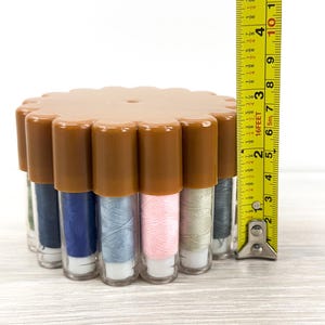 May include: A circular brown plastic container holding multiple vertical clear tubes of sewing thread in various colors, including blue, pink, and grey. A yellow measuring tape is placed vertically next to it, showing a height of 2.5 inches.