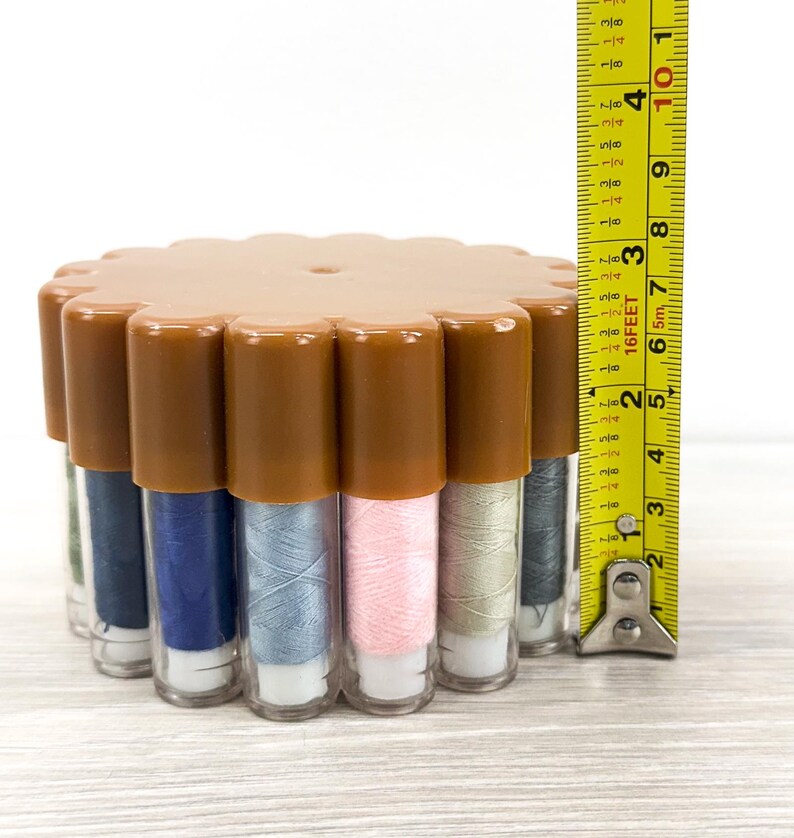 May include: A circular brown plastic container holding multiple vertical clear tubes of sewing thread in various colors, including blue, pink, and grey. A yellow measuring tape is placed vertically next to it, showing a height of 2.5 inches.
