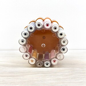 May include: A circular, transparent plastic organizer stand holding multiple cylindrical tubes. The stand features a brown base and a clear outer frame, with various colored cosmetic or craft tubes arranged vertically in a ring formation.