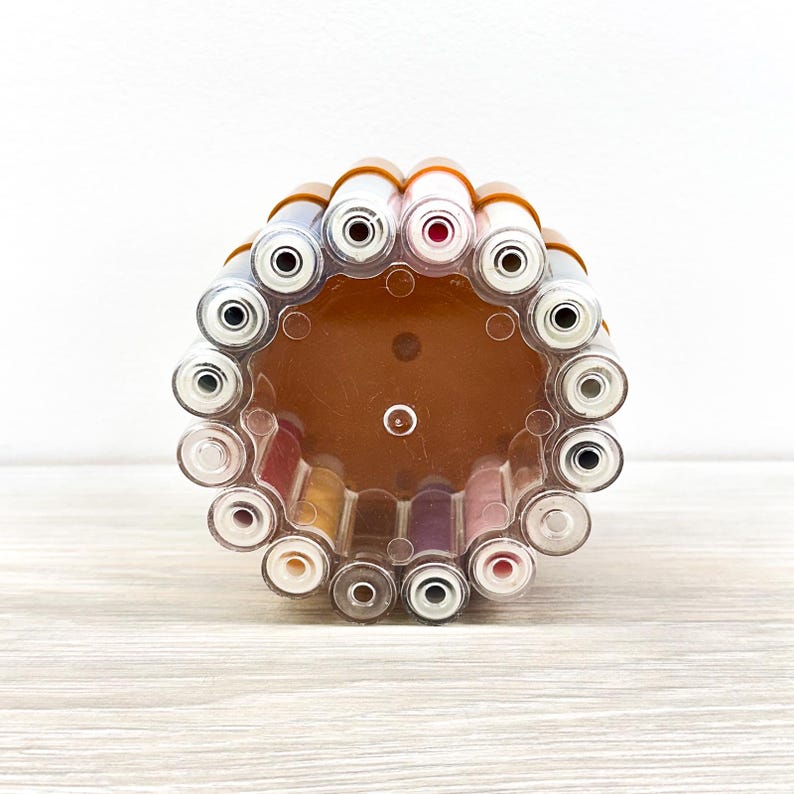 May include: A circular, transparent plastic organizer stand holding multiple cylindrical tubes. The stand features a brown base and a clear outer frame, with various colored cosmetic or craft tubes arranged vertically in a ring formation.
