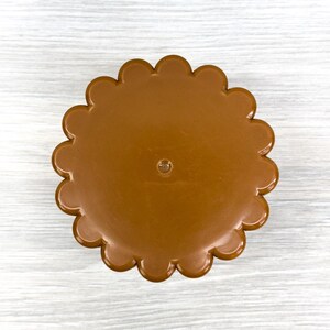 May include: A circular brown plastic lid featuring a scalloped edge design. The surface is smooth and solid in color with a small, centered circular indentation. The object is presented from a top-down perspective, highlighting its uniform shape.
