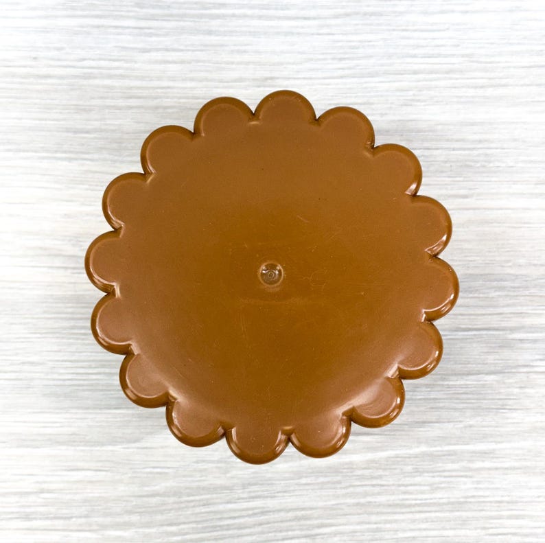 May include: A circular brown plastic lid featuring a scalloped edge design. The surface is smooth and solid in color with a small, centered circular indentation. The object is presented from a top-down perspective, highlighting its uniform shape.
