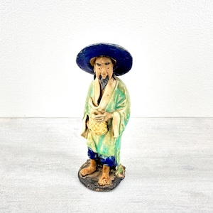 May include: A ceramic figurine of a person wearing a wide-brimmed dark blue hat, a light green robe with cream-colored lapels, and blue trousers. The figure has a goatee, holds a small yellow basket, and stands on a dark, textured base.