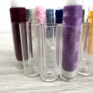 May include: A collection of cylindrical clear plastic holders containing various spools of sewing thread in colors including deep burgundy, light gray, soft pink, slate blue, dark navy, vibrant purple, and golden yellow arranged in a semi-circle.