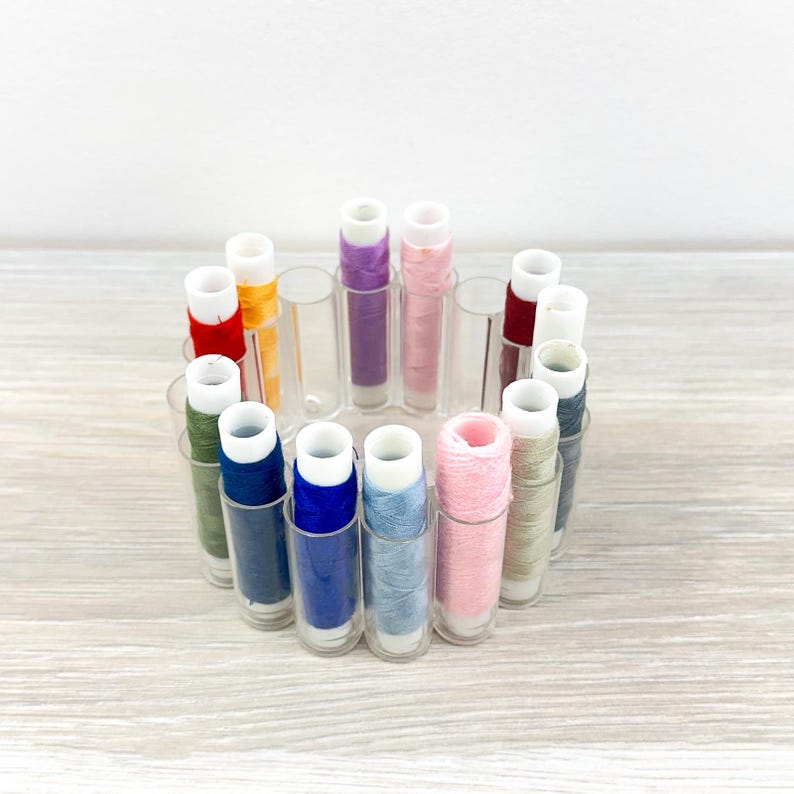 May include: A circular organizer holding multiple spools of sewing thread in various colors including red, yellow, purple, pink, blue, green, and grey. The organizer features individual clear plastic cylindrical slots for each spool of thread.
