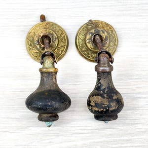 May include: Two matching drop-style drawer pulls featuring circular brass backplates with an intricate geometric pattern. Each pull has a dark, weathered, pear-shaped handle attached to a metal post, showing signs of age, patina, and worn black finish.