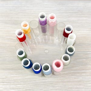 May include: A circular clear plastic thread spool holder containing twelve individual spools of sewing thread in various colors including red, orange, purple, pink, white, dark gray, light blue, royal blue, and olive green.