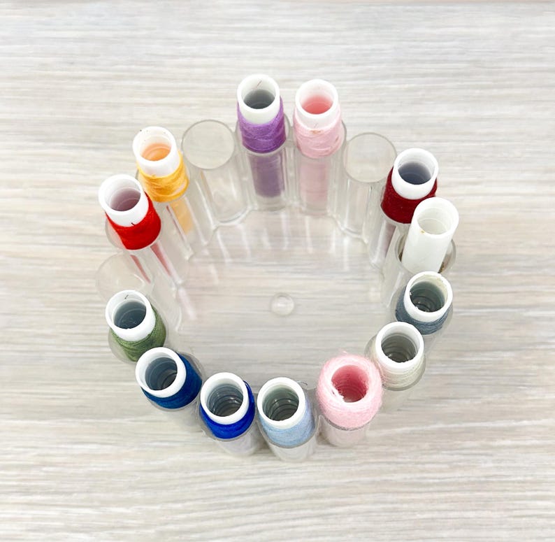 May include: A circular clear plastic thread spool holder containing twelve individual spools of sewing thread in various colors including red, orange, purple, pink, white, dark gray, light blue, royal blue, and olive green.