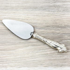 May include: A silver-toned cake server featuring a smooth, triangular metal blade and a decorative, ornate handle with intricate embossed floral and scroll patterns. The utensil has a polished finish and a tapered shape designed for slicing and serving.