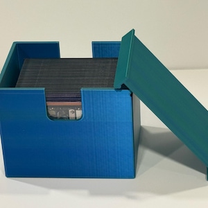 Può includere: Una scatola blu per riporre carte con un coperchio verde acqua. La scatola è aperta, rivelando una pila di carte all'interno. La scatola ha una forma rettangolare con un ritaglio su un lato per un facile accesso alle carte. Il coperchio è angolato.