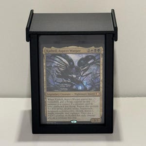 Può includere: Un espositore nero che contiene una carta di Magic: The Gathering. La carta presenta il testo "Kathril, Aspect Warper" e un'illustrazione di creatura. L'espositore ha un coperchio incernierato e un fronte trasparente.