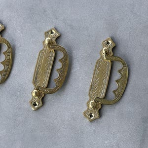 May include: Close-up of three antique brass drawer pulls. Each pull features an ornate, rectangular backplate with a curved handle. The brass has a warm, golden hue, and the design includes engraved floral patterns and decorative edges.