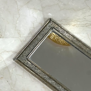 Può includere: Uno specchio rettangolare argentato con una cornice ornata, intricata e dettagliata. La cornice presenta un motivo geometrico.