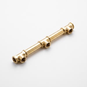 Op de afbeelding: Een messing sanitair verdeelstuk met een lange centrale buis verbonden aan drie T-stuk fittingen met schroefdraad. Het metalen onderdeel heeft een gladde, reflecterende afwerking en bevat meerdere ronde openingen met interne schroefdraad.