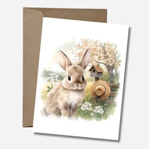 Peut inclure: Illustration à l'aquarelle d'un lapin assis dans un champ avec des marguerites, un nichoir et un chapeau de paille. L'arrière-plan présente un arbre en fleurs et une clôture en bois. La carte est posée sur une enveloppe marron.