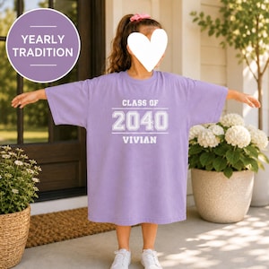May include: A light purple short-sleeved t-shirt featuring white screen-printed text that reads CLASS OF 2040 VIVIAN. The shirt has a crew neckline and a loose, relaxed fit. A circular purple badge in the corner displays the text YEARLY TRADITION.
