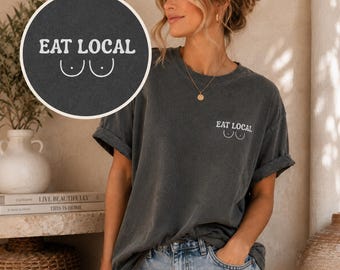 May include: A charcoal grey short-sleeve t-shirt featuring a white graphic on the left chest that reads EAT LOCAL above a simple line drawing of two breasts. The shirt has a crew neckline and a relaxed fit, paired with light blue denim jeans.