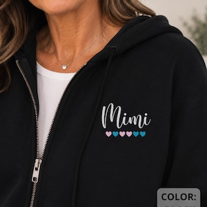 May include: A black zip-up hoodie featuring the white script text Mimi on the left chest, positioned above a row of five small hearts alternating in light pink and light blue colours. The garment includes a metal zip and a hood with drawstrings.
