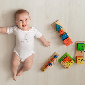 May include: A baby wearing a white onesie with the text "what you do makes a difference" lies on a light wood floor.  The baby is smiling and looking up at the camera.  There are four wooden toys on the floor, including a shape sorter, a bead maze, a stacking toy, and a toy with a string and beads.