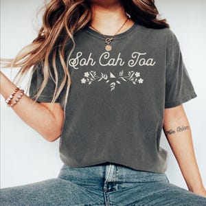 May include: A dark gray t-shirt with the text "Soh Cah Toa" in white lettering with a floral design underneath.