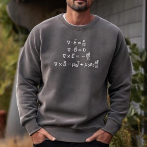 Könnte beinhalten: Ein dunkelgraues Sweatshirt mit weißem Text, der die Maxwellschen Gleichungen des Elektromagnetismus zeigt. Die Gleichungen lauten: ∇ · E = ρ/ε₀, ∇ · B = 0, ∇ × E = -∂B/∂t, ∇ × B = μ₀J + μ₀ε₀∂E/∂t.