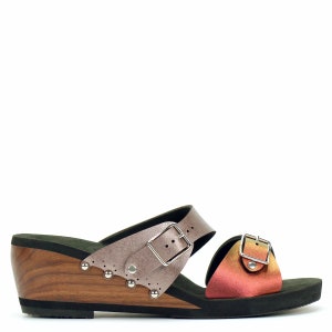 Op de afbeelding: Een paar bruine en zilveren wedgesandalen met twee banden en gespen. De banden zijn versierd met zilveren studs. De sandalen hebben een houten wedge hak en een zwarte zool.