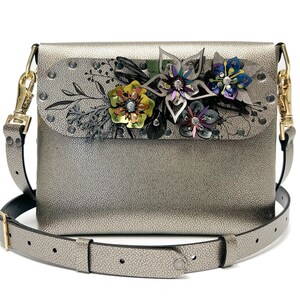 May include: A silver crossbody bag with a flap closure and a floral embellishment. The flowers are made of leather and have a variety of colors, including green, yellow, pink, and purple. The bag has a black adjustable strap.