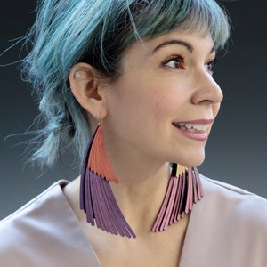 May include: A woman with blue hair wearing long, layered leather earrings. The earrings are a mix of pink, purple, and orange.