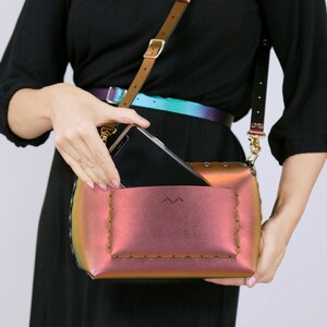 May include: A small, iridescent pink crossbody bag with a flap closure and a stitched detail around the edges. A black phone is partially visible inside the bag.