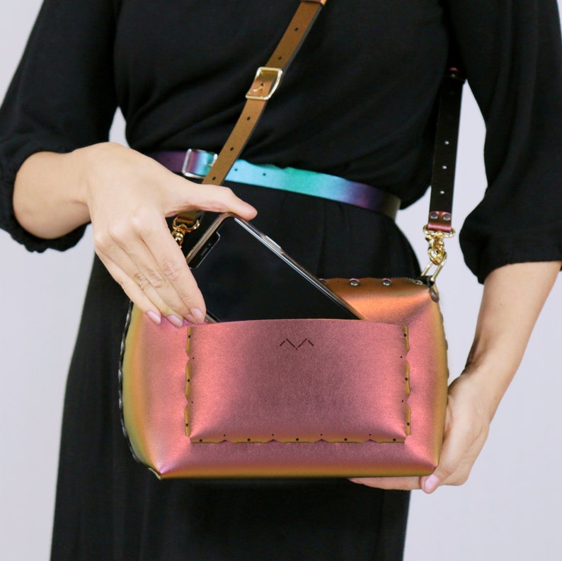 May include: A small, iridescent pink crossbody bag with a flap closure and a stitched detail around the edges. A black phone is partially visible inside the bag.
