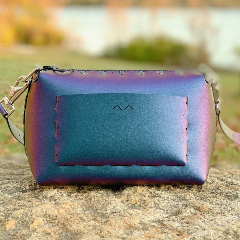 May include: A rectangular iridescent crossbody bag with a flap pocket. The bag has a zipper closure and a silver-colored strap. The bag's color shifts between purple, blue, and green. The flap pocket has a decorative edge and a small logo.