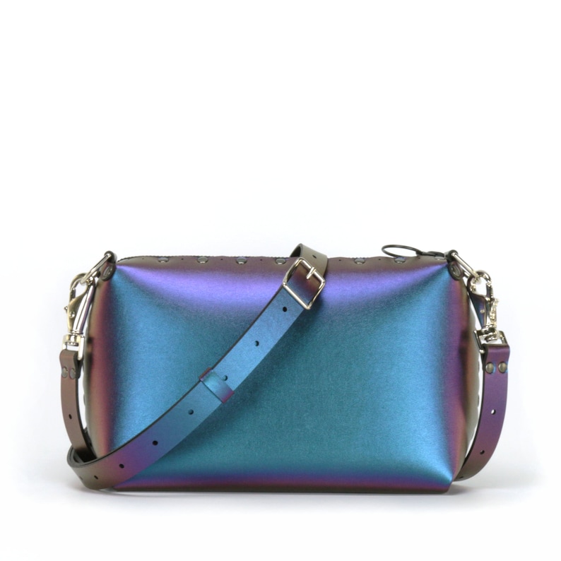 May include: Iridescent rectangular handbag with a shoulder strap and silver-tone hardware. The bag transitions from purple to teal and blue. The strap is black with a silver buckle. Small silver rivets line the top of the bag.