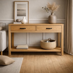 Op de afbeelding: Een lichte eikenhouten console met twee lades en een onderste plank. De tafel heeft zwarte handgrepen en is gestyled met een ingelijste landschapsprint, vazen en een rieten mand. De kamer heeft houten vloeren en een neutraal kleurenpalet.