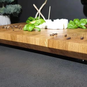 May include: A large, rectangular wooden serving board with a natural wood finish. The board has a beveled edge and is sitting on a dark gray surface. There are small black peppercorns scattered on the board, along with some white cheese and green herbs.
