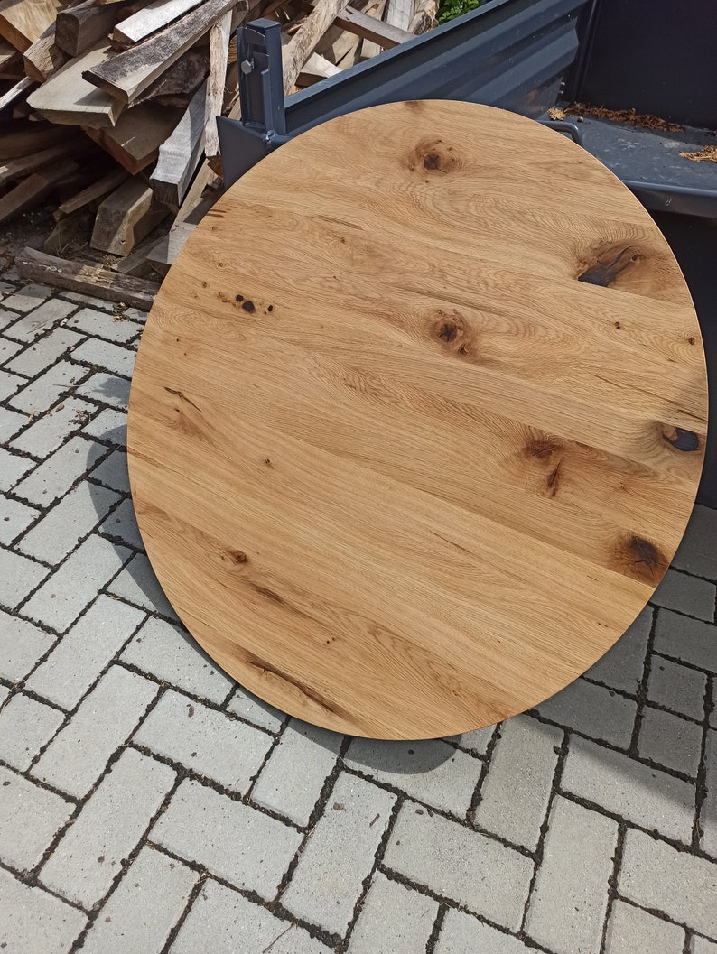 以下が含まれることがあります： 自然な木目仕上げの丸い木製テーブルトップ。テーブルトップは薄い茶色で、濃い茶色の節と木目が特徴です。テーブルトップは灰色のレンガの表面の上に置かれています。