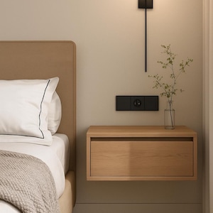 Puede incluir: Un dormitorio moderno con una mesita de noche flotante de madera con un cajón. Encima, una lámpara de pared negra con una bombilla esférica. Se ve una cama con cabecero beige y ropa de cama blanca.