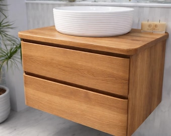 May include: A wall-mounted, two-drawer wooden vanity with a light oak finish. A white, round, ribbed ceramic sink sits atop the vanity. A small candle and gold faucet are visible in the background. The vanity is designed for a bathroom.