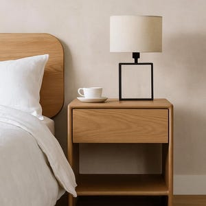 Op de afbeelding: Een houten nachtkastje met een lade en een plank. Een zwarte lamp met vierkant frame en een beige lampenkap staat erop. Een witte kop en schotel staan ook op het nachtkastje. Een bed met wit beddengoed is gedeeltelijk zichtbaar.