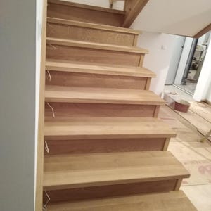 Puede incluir: Una escalera de madera con tonos de madera natural. Los peldaños tienen un acabado marrón claro y están hechos de madera maciza. La escalera está en una casa o edificio en construcción.