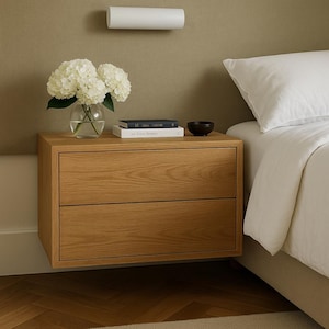 May include: A two-drawer wooden bedside table with a light oak finish. A glass vase with white flowers, books, and a small black bowl sit on top. A white wall-mounted light fixture is above the table, and a bed with white linens is to the right.