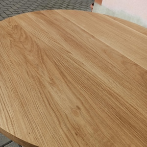 May include: A close-up of a round wooden table top with a natural wood grain finish. The table top is made of solid wood and has a smooth, polished surface.