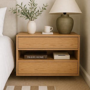 May include: A light oak floating bedside table with two drawers and an open shelf. A white vase with greenery, a mug, and a lamp with a beige shade sit on top. A book with the text "TRAVEL HOME" is on the shelf.