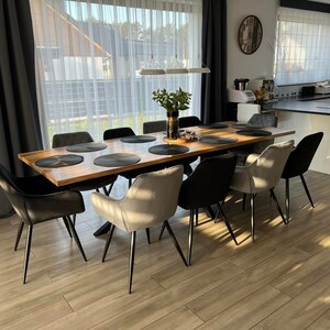 Può includere: Un tavolo da pranzo in legno con gambe in metallo nero e otto sedie. Le sedie sono rivestite in tessuto nero e grigio. Il tavolo è apparecchiato con sottopiatti e un vaso di verde.