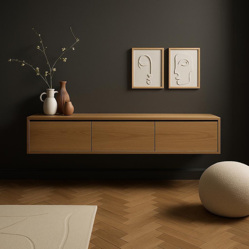 May include: A light brown, floating wooden cabinet with three drawers. Above the cabinet are two framed, minimalist face art pieces. On the cabinet's surface are three vases and a sprig of flowers. A cream-colored rug and a round, fuzzy ottoman are in the foreground.