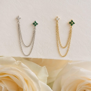 May include: Two double-chain earrings, one silver-toned and one gold-toned. Each earring features a single clear stone stud connected by two delicate chains to a four-petaled flower-shaped stud set with four small green stones.