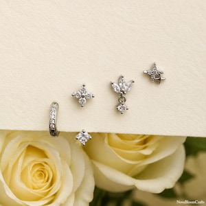 May include: A set of five silver-toned earrings featuring clear cubic zirconia stones. The collection includes a small hoop, a four-petaled flower stud, a marquise-leaf drop stud, a single round stud, and a four-pointed star stud with a small accent.