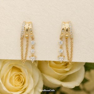 May include: A pair of gold-toned earrings featuring a triple-banded top design accented with small clear stones. Each earring includes a delicate gold chain and a vertical drop strand ending in a triangular clear stone, with NovaBloomCrafts text.