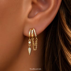 May include: Three gold hoop earrings connected by chains. The first hoop features a dangling charm with a round stone and a pointed, stone-encrusted tip. The second hoop is plain. The third hoop is lined with small clear stones and a double chain.