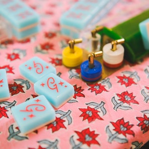 May include: Four blue and white tiles with red stars and the letter "G" on a pink floral background. The tiles are used for a game like Mahjong.