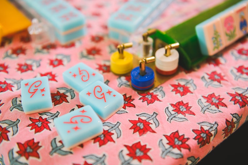 May include: Four blue and white tiles with red stars and the letter "G" on a pink floral background. The tiles are used for a game like Mahjong.